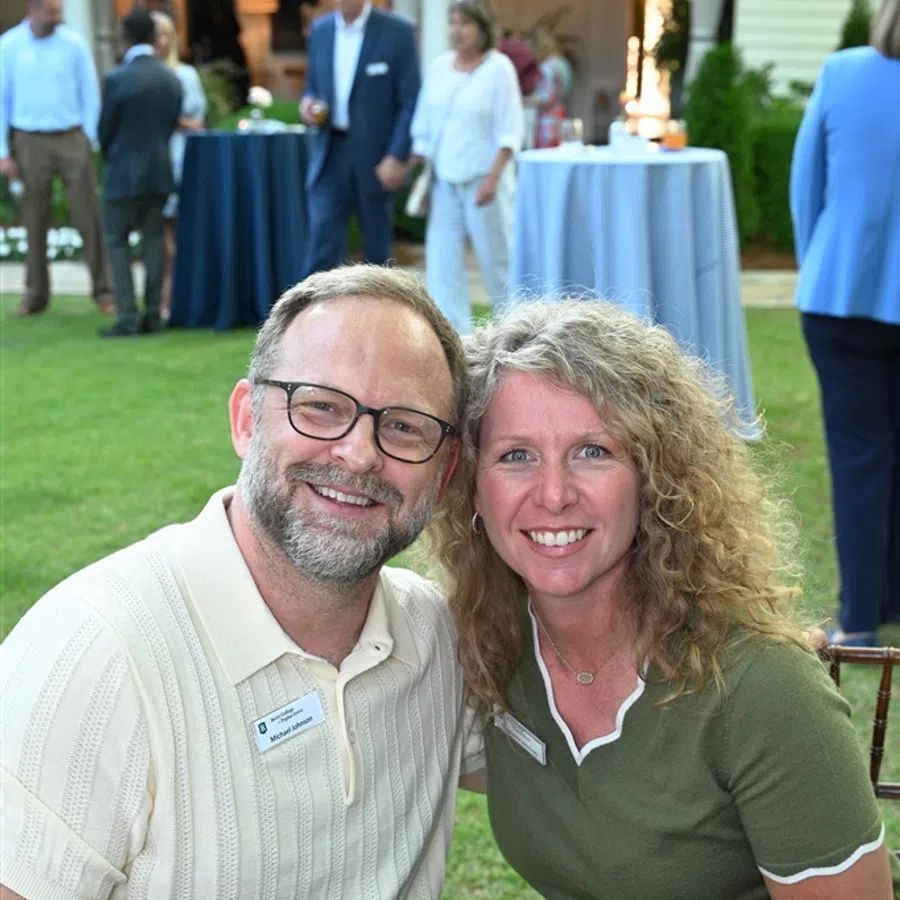Smiling couple posing at outdoor event with people and tables in the background on green lawn.