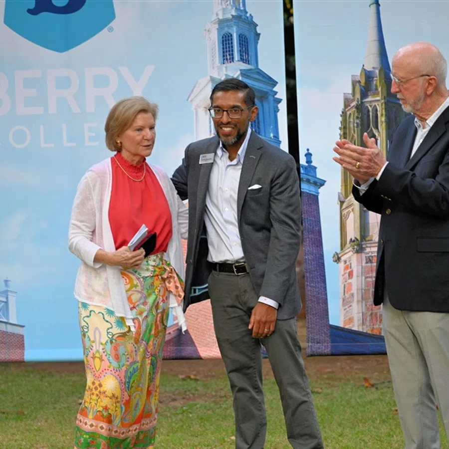 Three people at Berry College event with campus buildings backdrop, one man clapping and two smiling individuals.