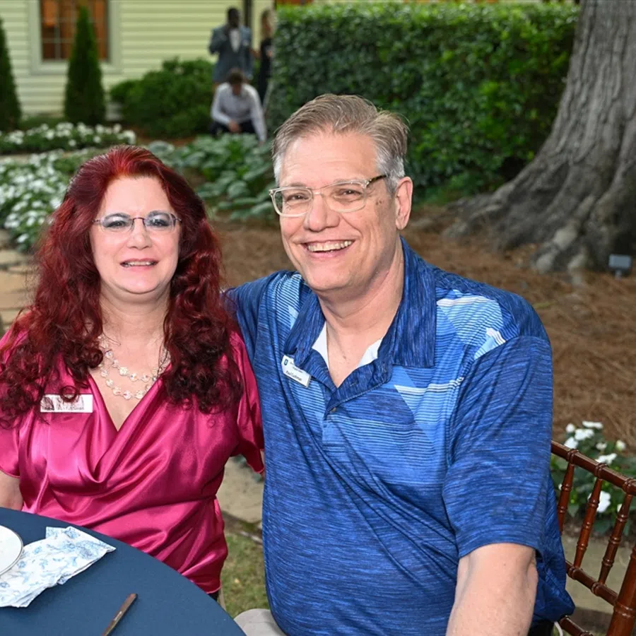 Smiling middle-aged couple seated at outdoor table during garden event with greenery and house in background