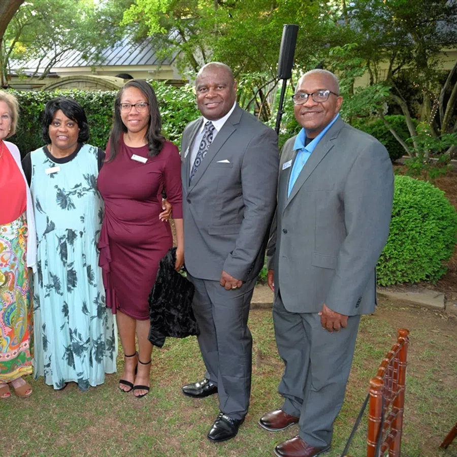 Six diverse adults standing outdoors smiling, dressed in semi-formal attire at a garden event under trees.