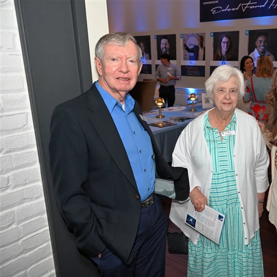 Three people smiling at a social event with portraits displayed on the wall and attendees mingling in the background.