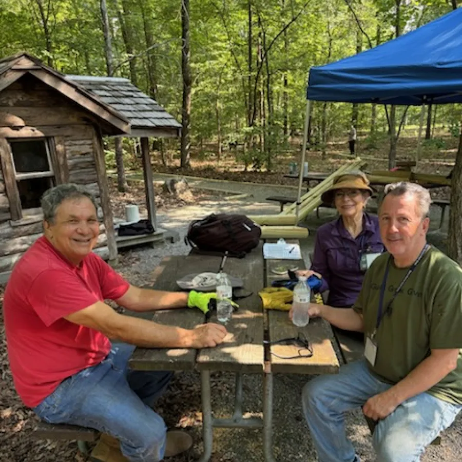 Three people sitting at a wooden picnic table in a forest park enjoying a break with water bottles and gear nearby.