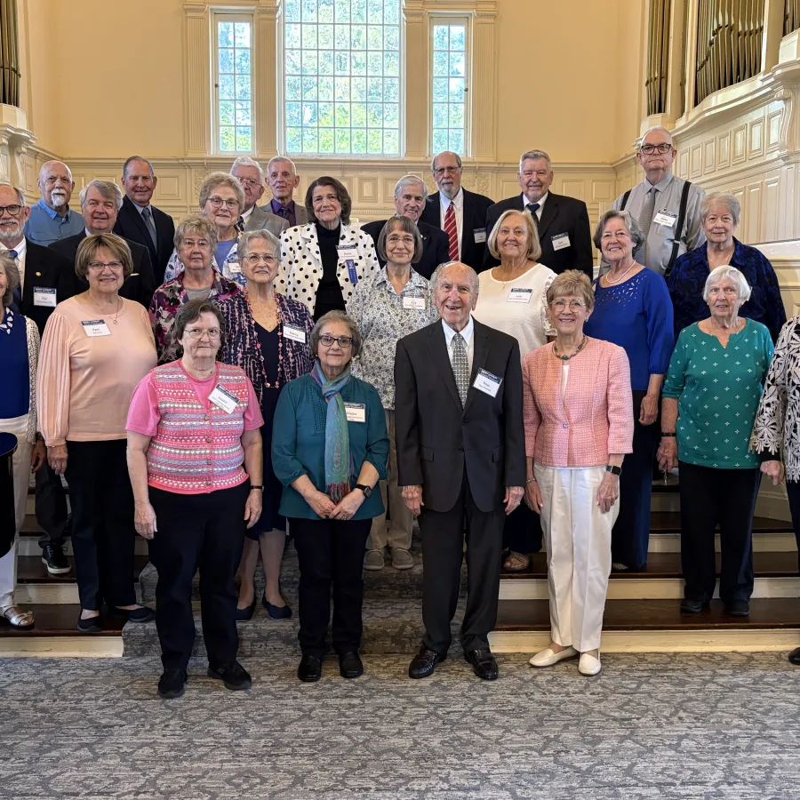 Group of 26 elderly adults posing in formal attire inside a church or auditorium with large windows.