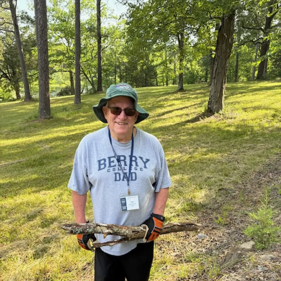 Smiling man wearing Berry College Dad shirt and gloves carrying tree branches in sunny wooded park.