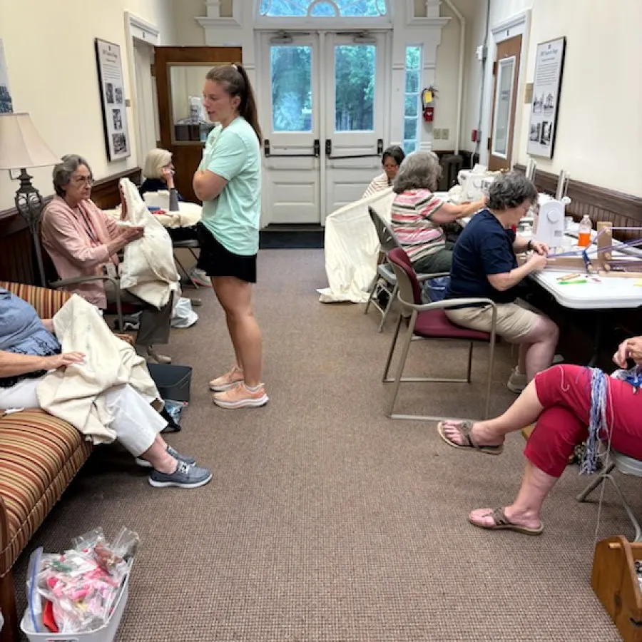Group of women sewing and knitting together in a bright community room with large arched window and neutral decor.