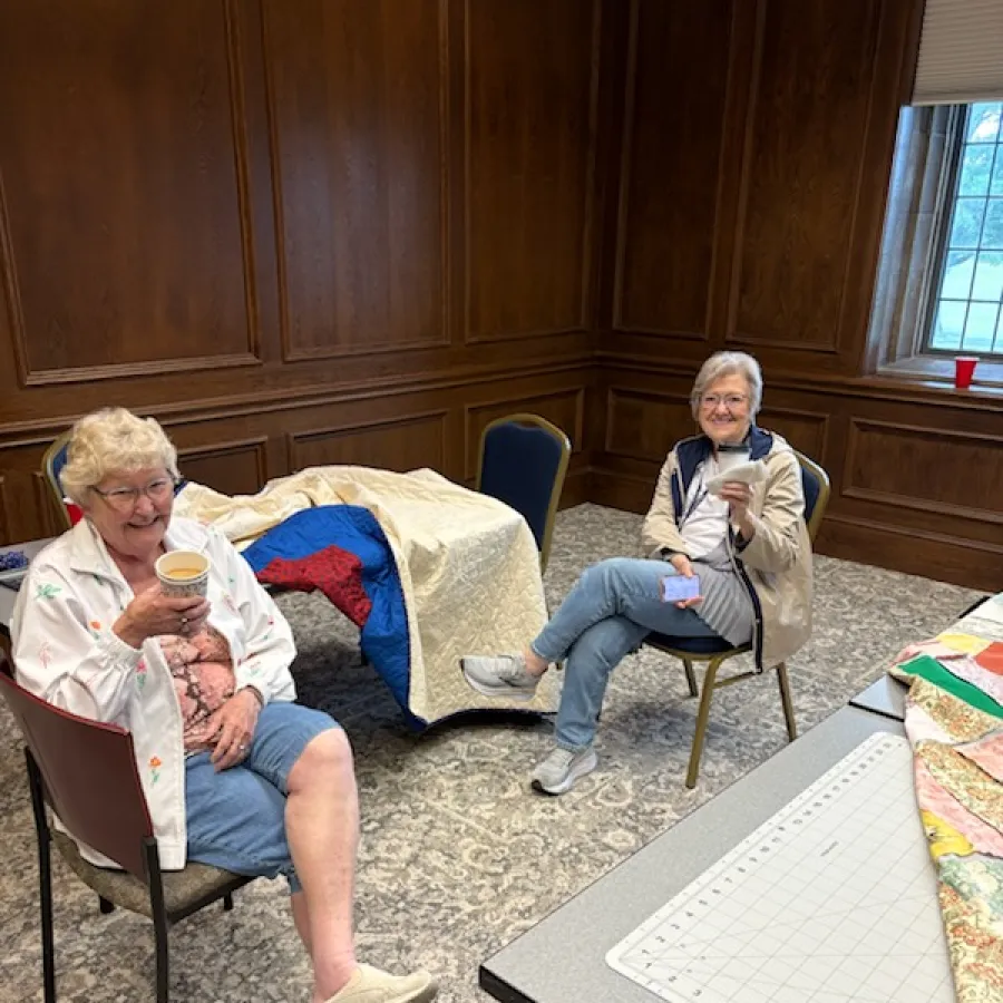 Two elderly women sitting in a cozy room with quilts, drinking coffee and smiling at the camera.