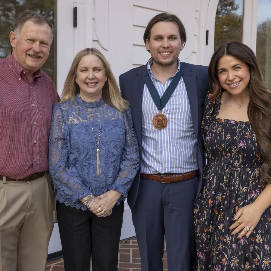 Four people smiling outside by a white building door, one wearing a medal, dressed in semi-formal attire.