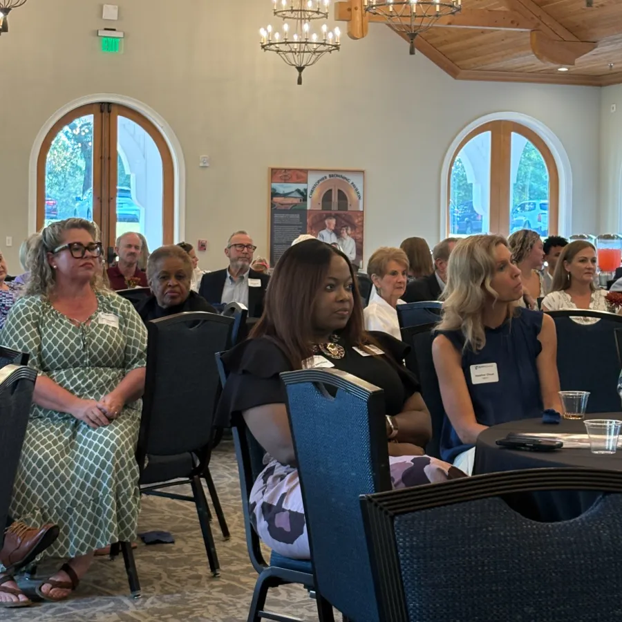 Diverse group of people seated and attentively listening in a bright room with wooden beams and chandeliers.