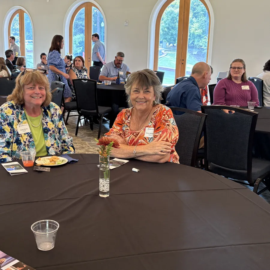 Two women smiling at a round table during a social event in a bright room with arched windows.