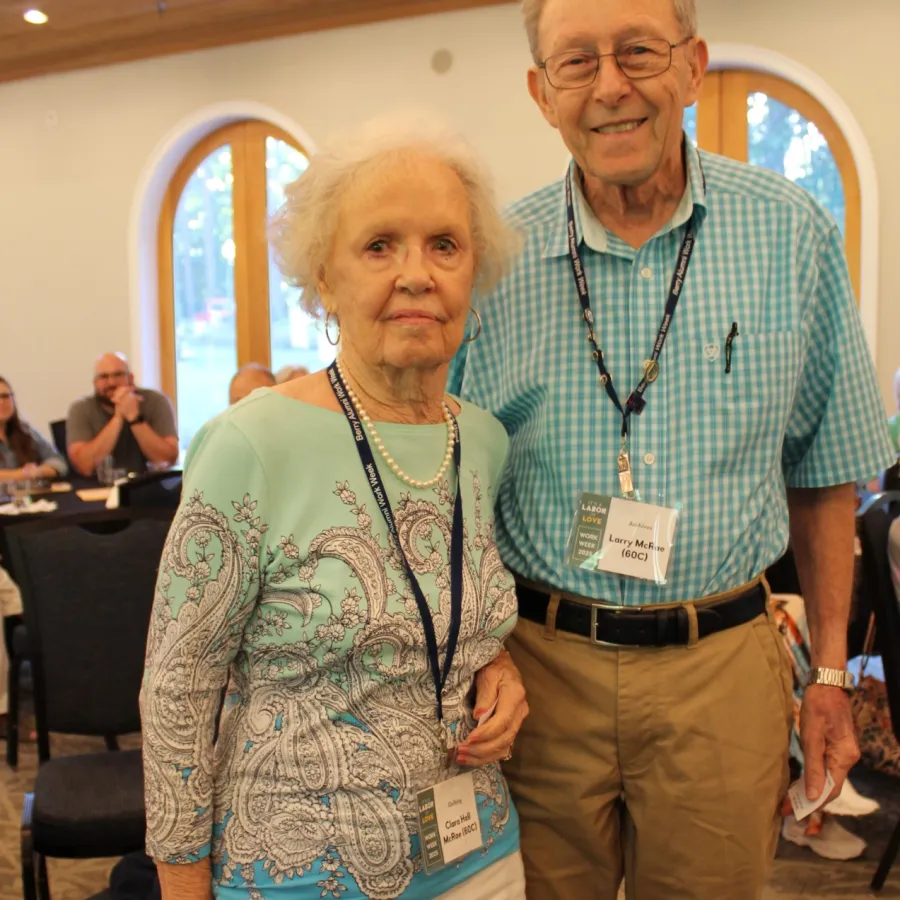 Elderly couple wearing name tags standing together in a bright room with wooden ceiling and large windows.