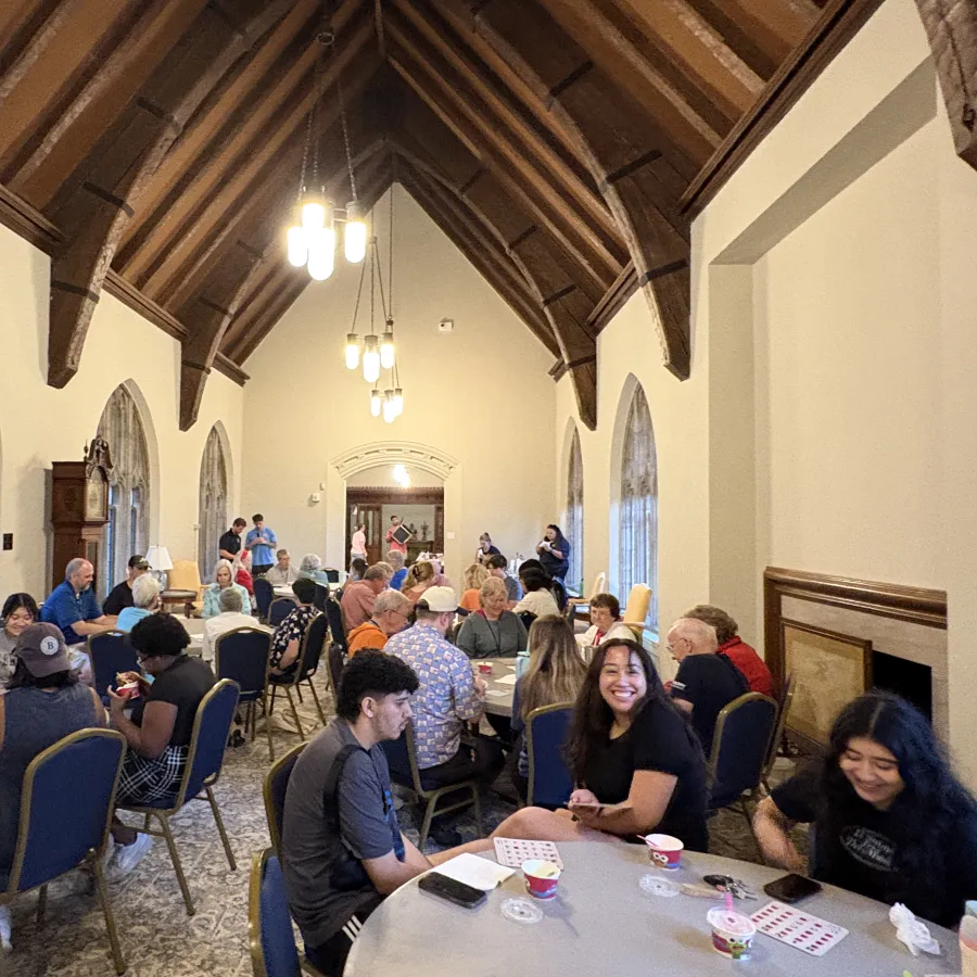 People gathered in a high-ceiling room with arched windows and chandeliers, enjoying a social event with tables and chairs.