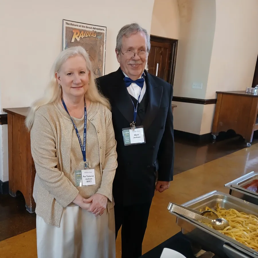 Two people wearing name badges stand near a buffet table with pasta in a formal room with wood furniture.