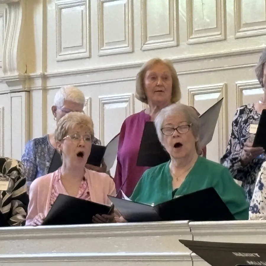 Senior women singing together in a choir, holding black folders in a bright, elegant room with white paneling.