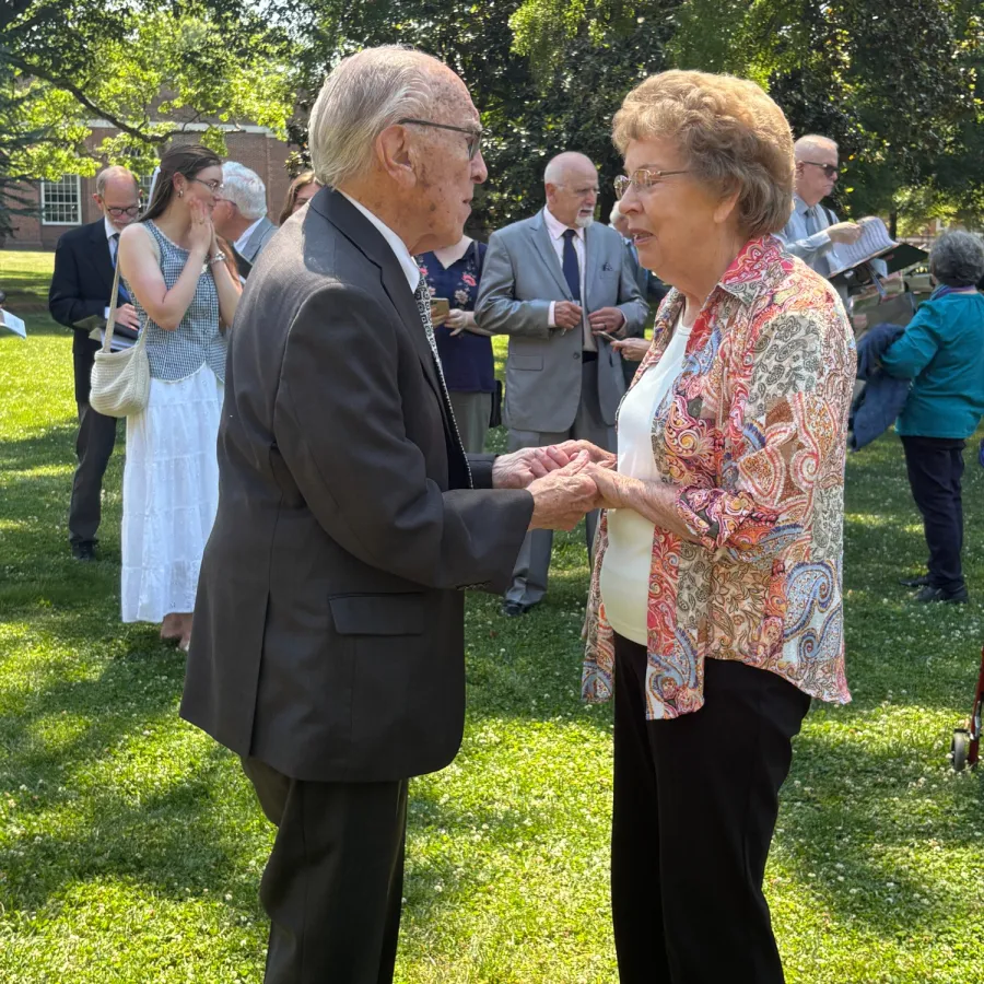 Elderly couple holding hands and talking outdoors in a sunlit garden setting with people socializing in the background