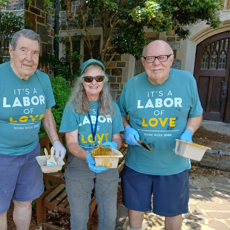 Three seniors wearing matching 'It's a Labor of Love' shirts volunteering outdoors with paintbrushes and gloves.