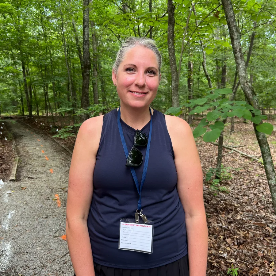 Smiling woman with silver hair in navy tank top standing on wooded trail with event badge and sunglasses.