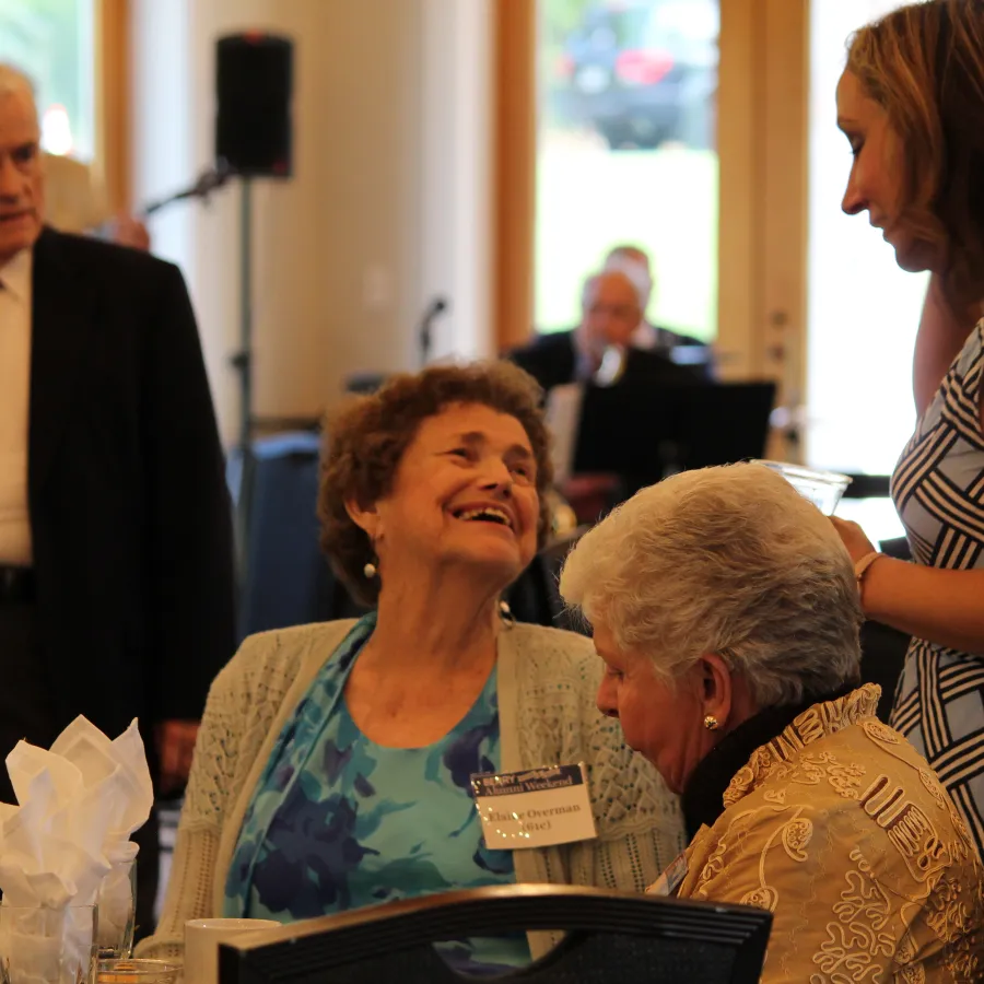 Three women and one man engaged in conversation at an indoor social event with tables and drinks.