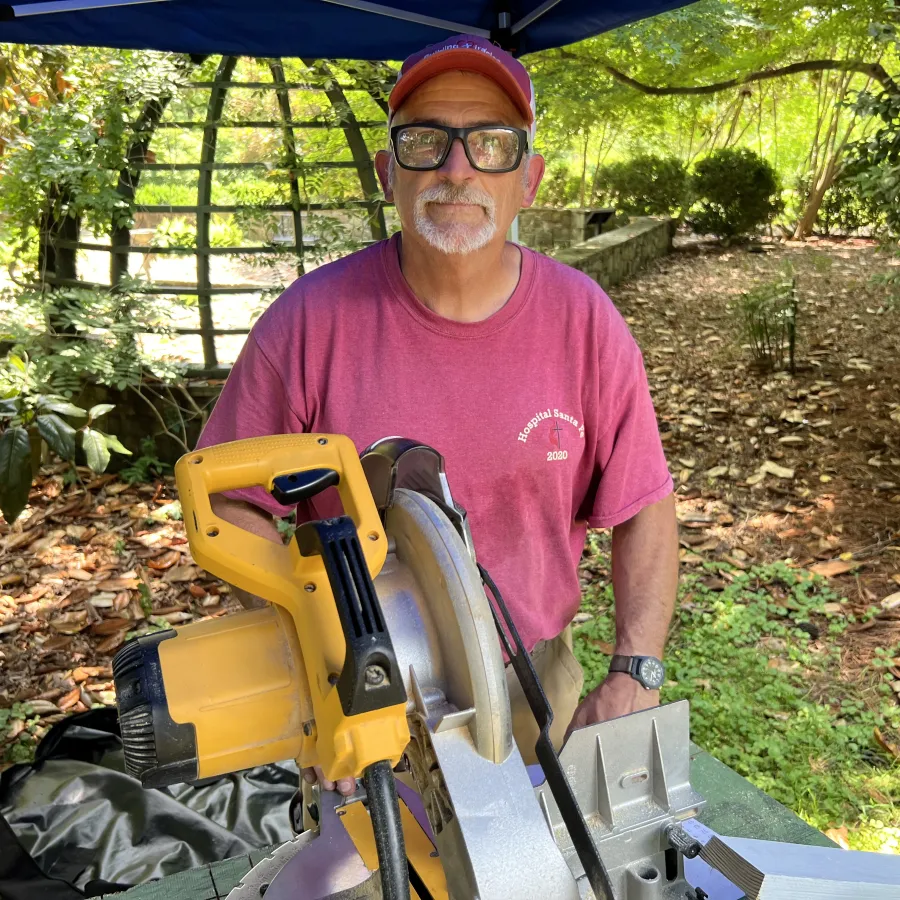 Man in red shirt and cap stands behind a large yellow circular saw outdoors surrounded by greenery.