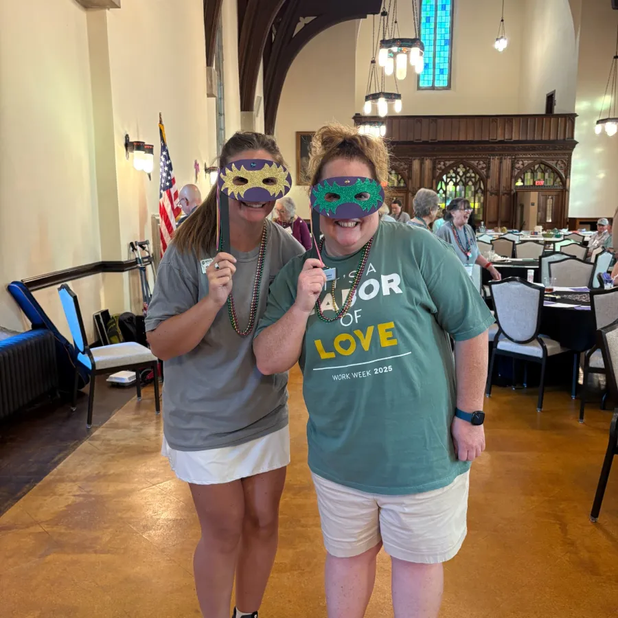 Two women in casual shirts and shorts wearing colorful masks and beads in a decorated room with flags and tables.