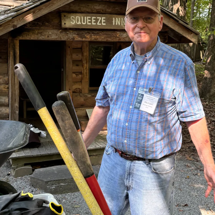 Man in a plaid shirt and cap stands outside a rustic wooden building labeled Squeeze Inn holding colorful paddles.