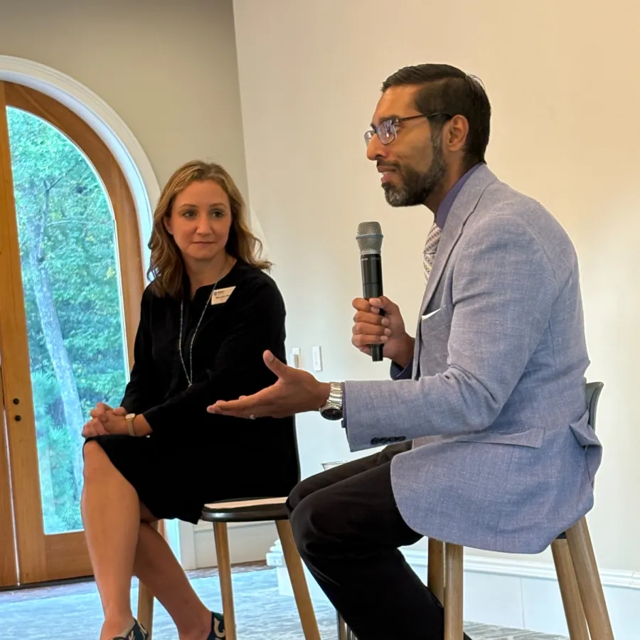 Man in gray jacket speaking into microphone seated next to woman in black dress sitting on stools indoors