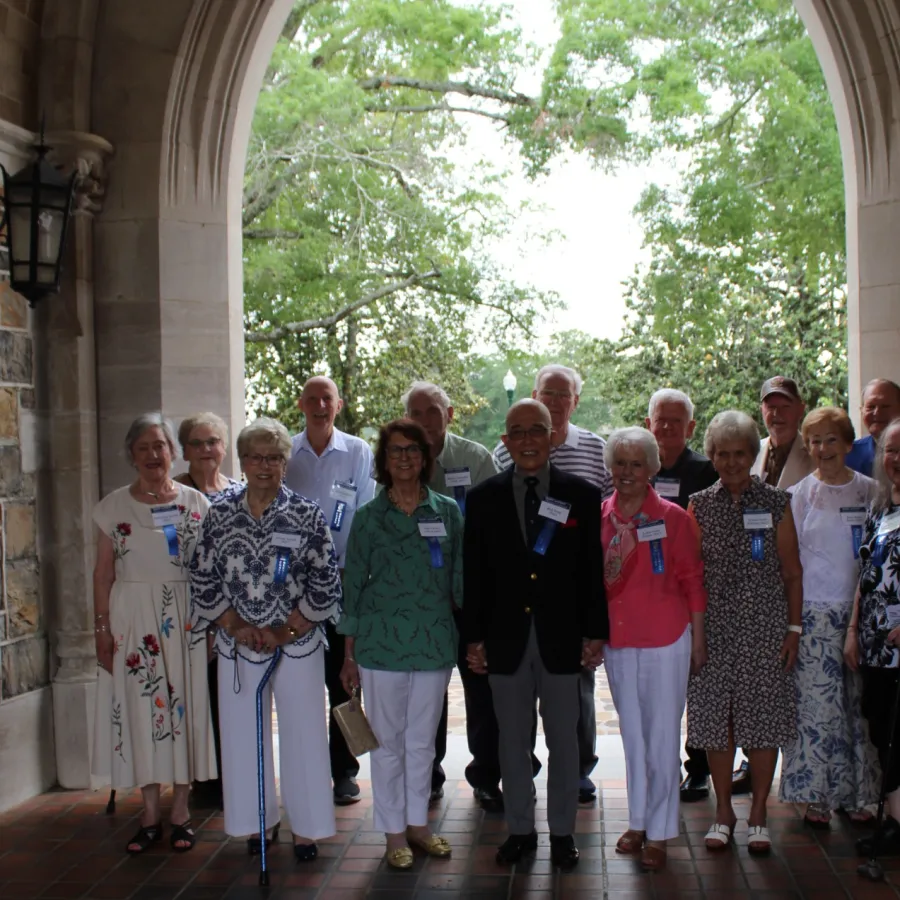 Group of elderly people standing in an arched outdoor corridor with trees visible through the arch.