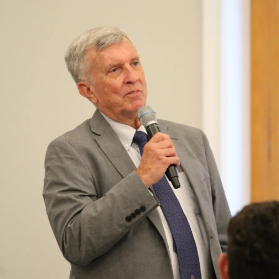 Senior man in gray suit speaking into microphone during indoor event or presentation.