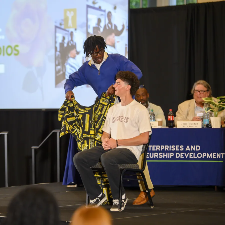 Person draping patterned fabric over seated man on stage during entrepreneurship event with panel in background