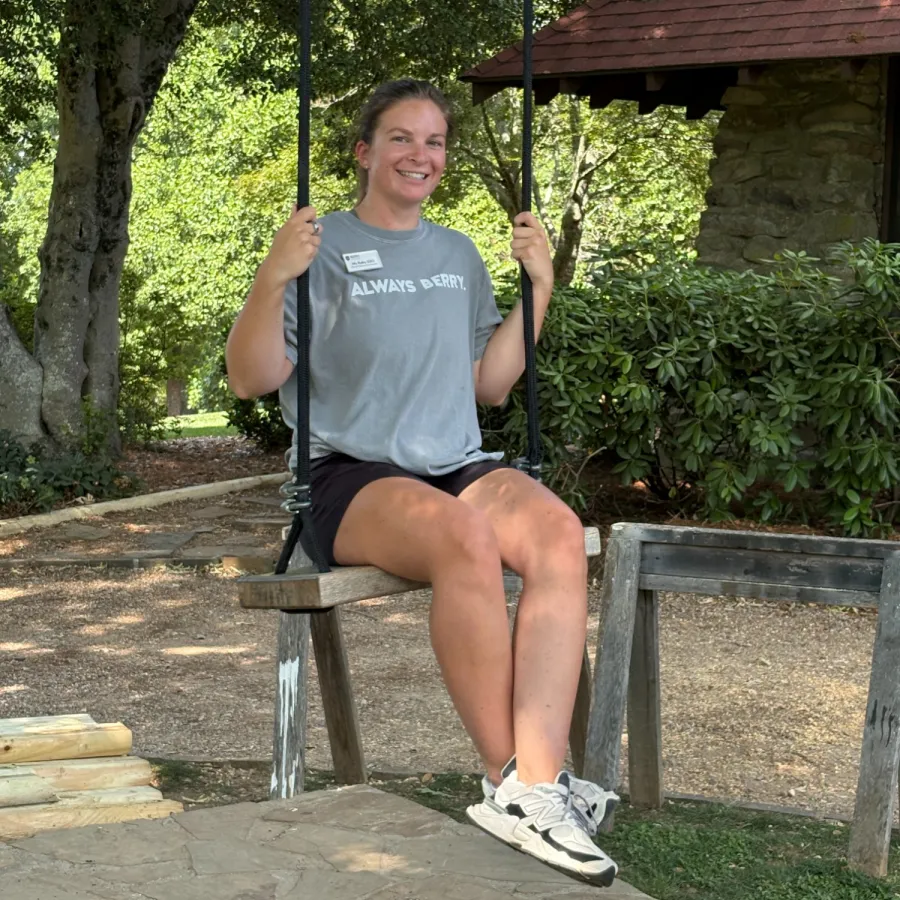 Woman in gray shirt and black shorts smiling on a wooden swing in a sunny garden with trees and stone path.