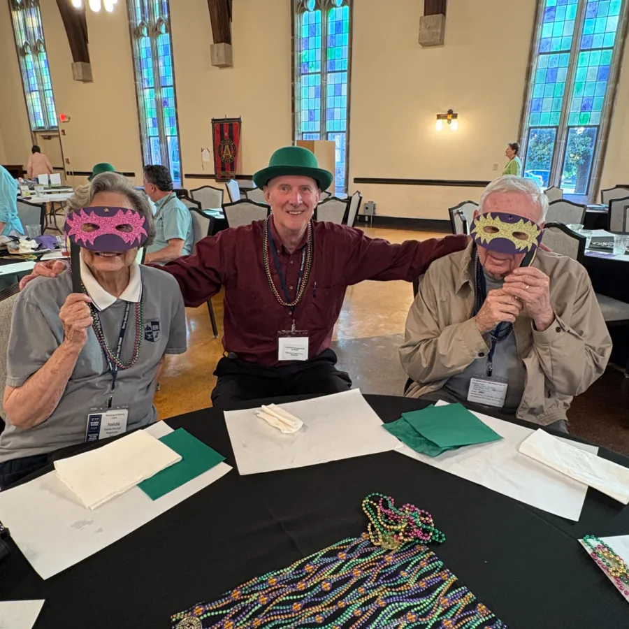 Three elderly people wearing festive masks and beads at a table with colorful Mardi Gras beads inside a church hall.