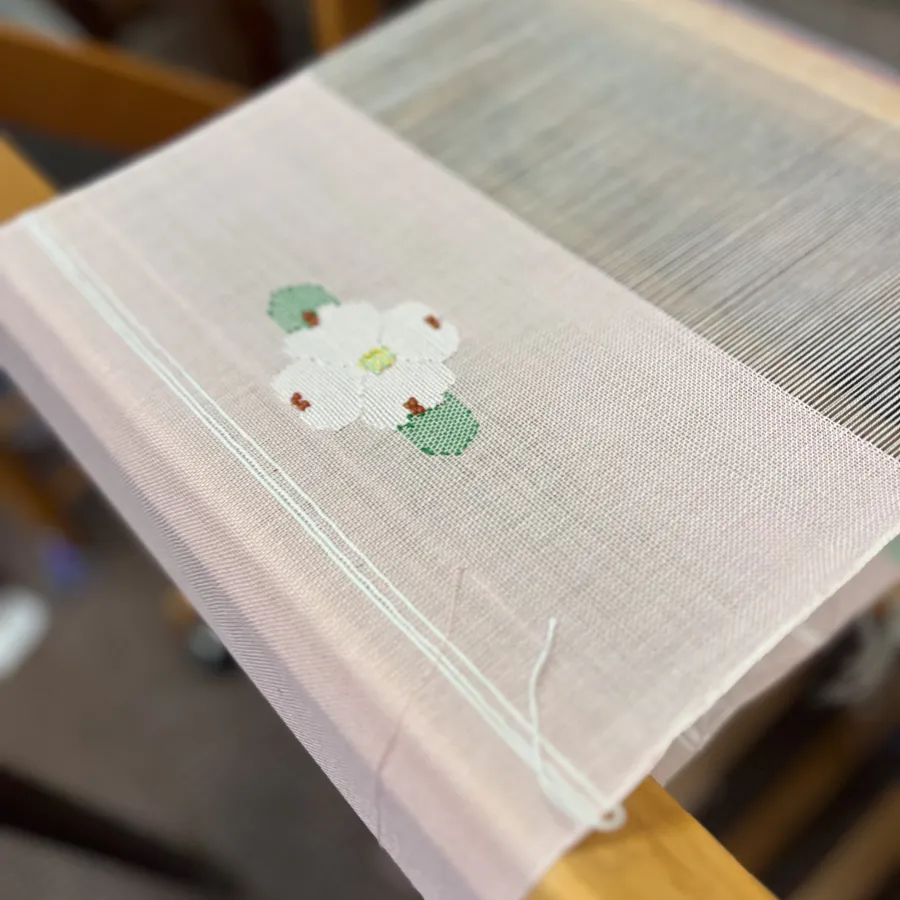 Close-up of a weaving loom with light pink fabric and a white flower with green leaves pattern in progress.