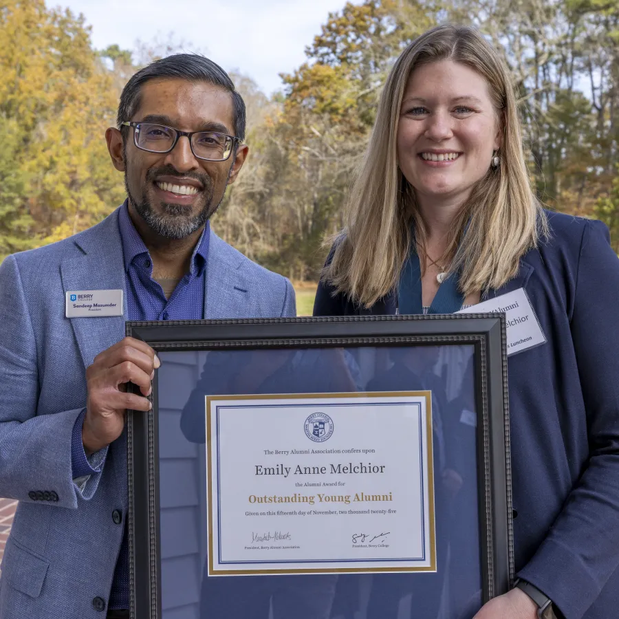 Two professionals outdoors smiling and holding a framed Outstanding Young Alumni certificate awarded to Emily Anne Melchior.