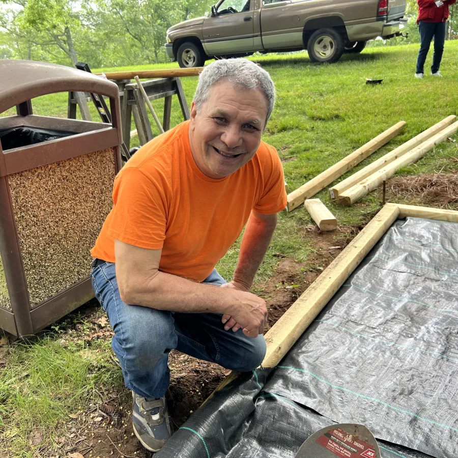 Man in orange shirt kneeling on grass next to wooden frame and black tarp near park trash can and trees.