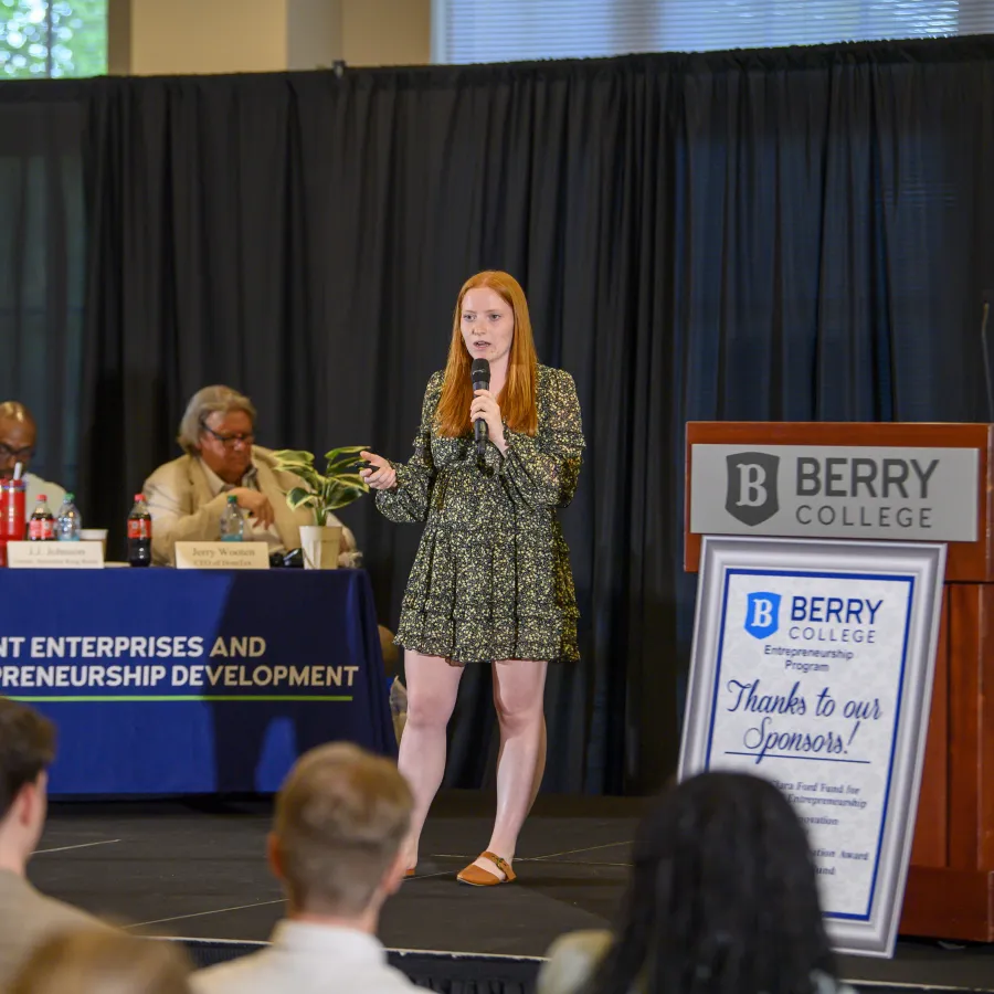 Young woman presenting with microphone at Berry College student entrepreneurship event with panel and audience.