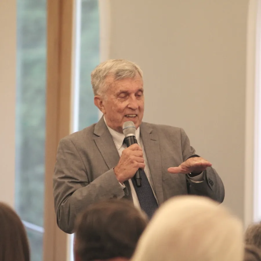 Older man in a suit speaking into a microphone during a presentation or speech at an indoor event.