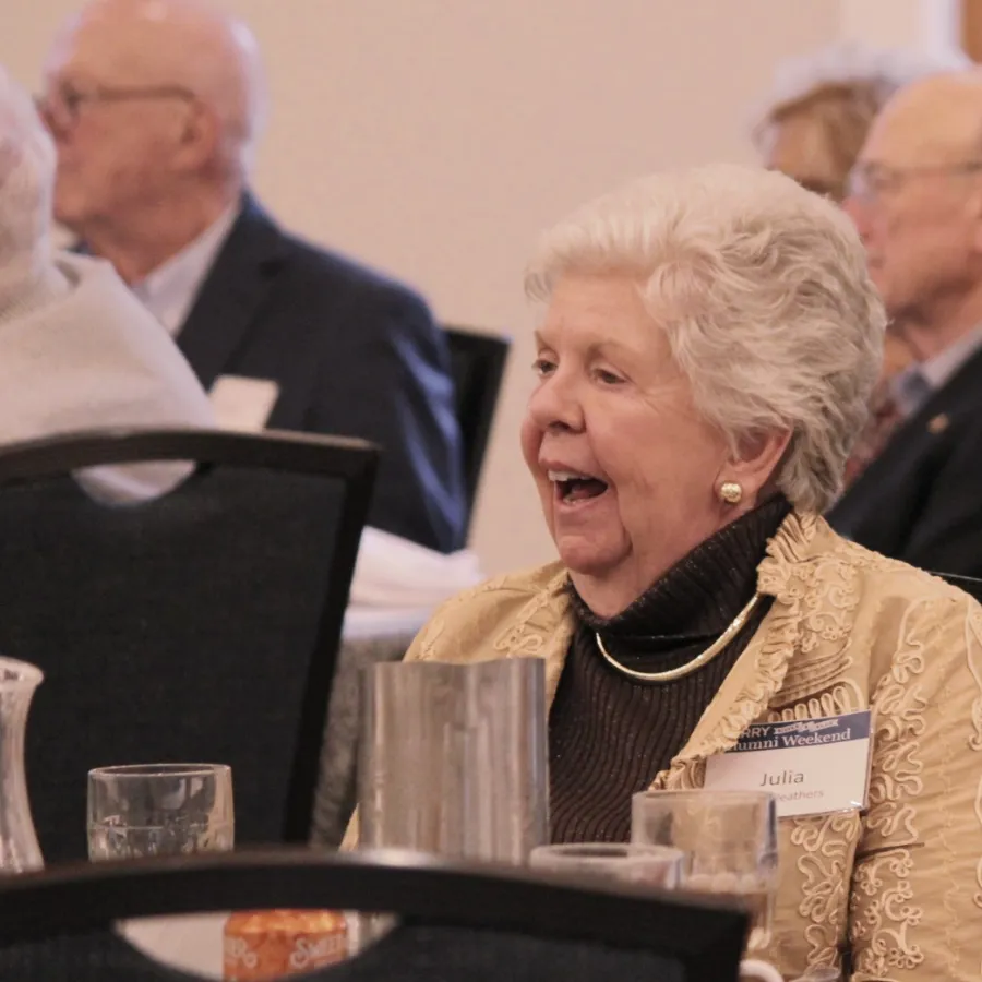 Elderly woman smiling and engaging in conversation at a social event with other seniors seated around tables.