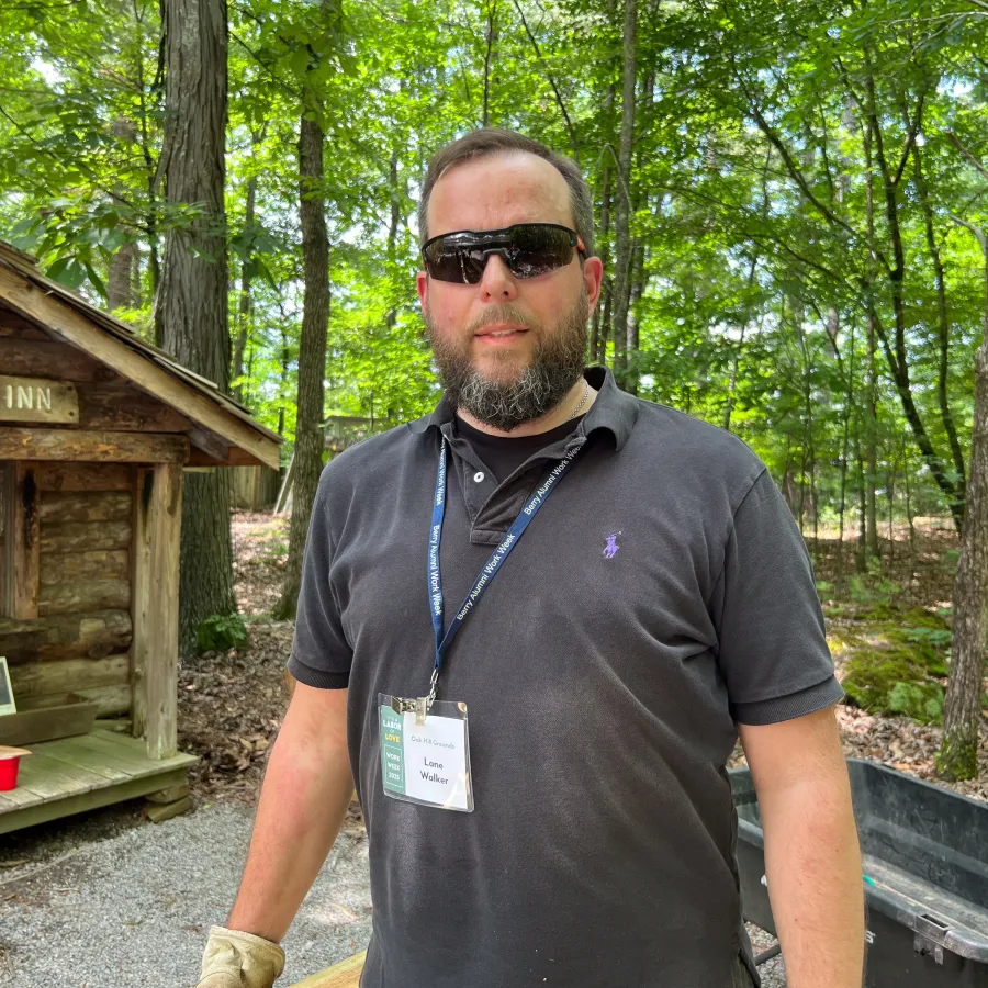 Bearded man with sunglasses wearing a black polo and badge stands outdoors near a wooden cabin in a forest.