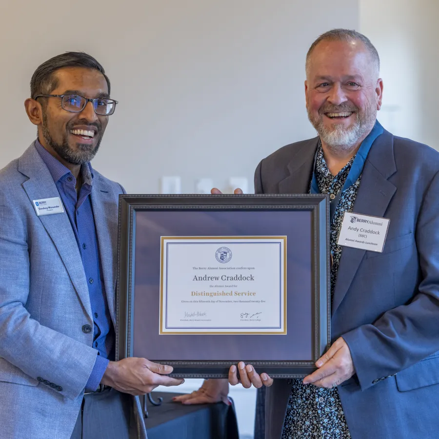 Two men smiling and holding a framed certificate of distinguished service awarded to Andrew Craddock indoors.