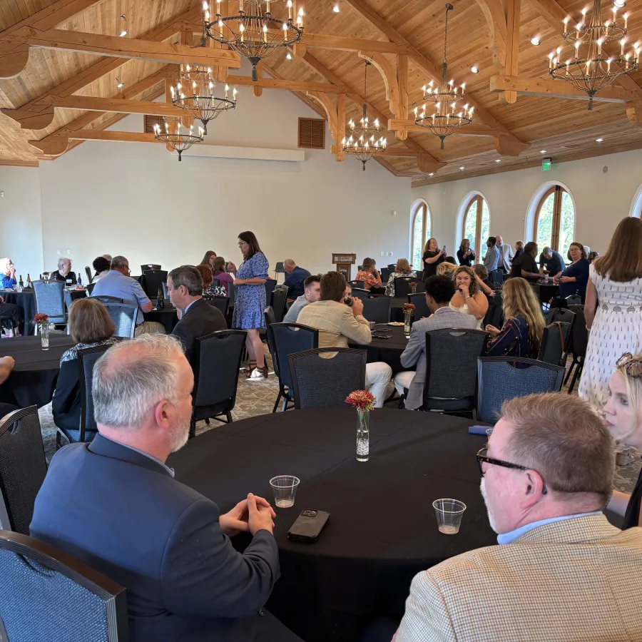 Guests socialize in a bright, wooden-ceiling event hall with round tables covered in black tablecloths.
