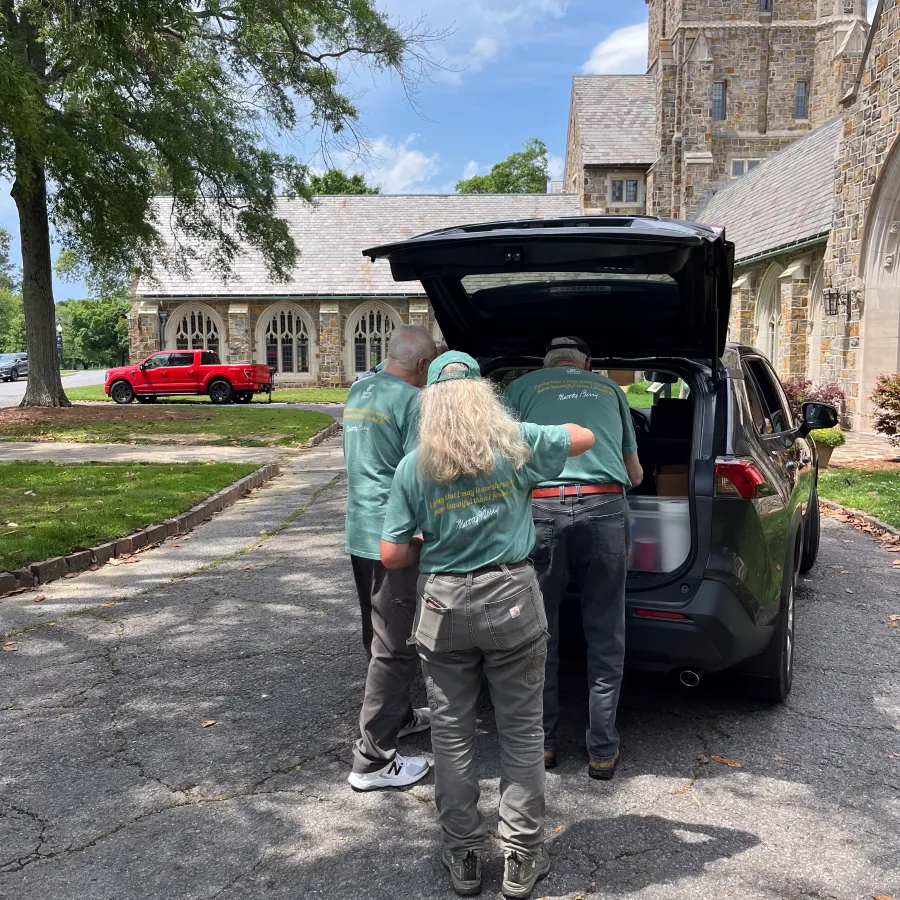 Three people in matching green shirts unload items from the back of a black SUV at a historic stone building.