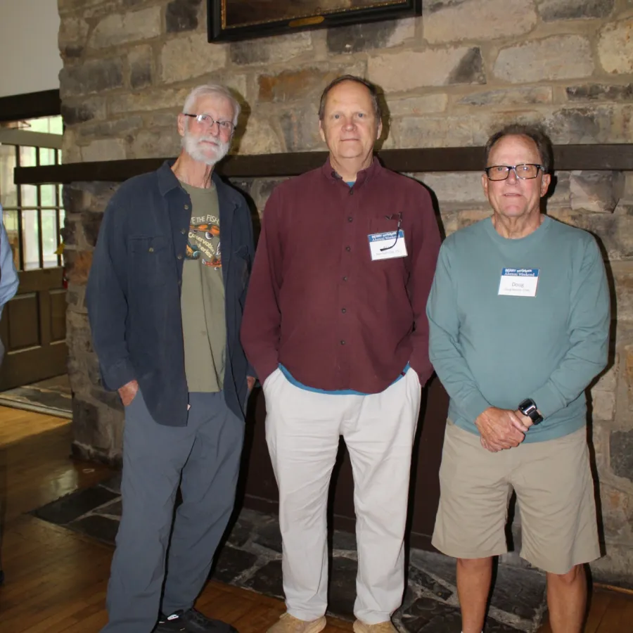 Three older men standing indoors on a wooden floor in front of a stone wall with a framed portrait hanging.