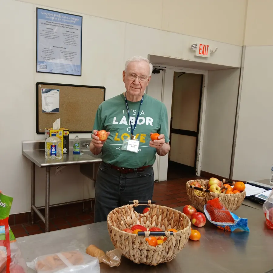 Elderly man holding apples and oranges in a kitchen with baskets of fruit on the table.
