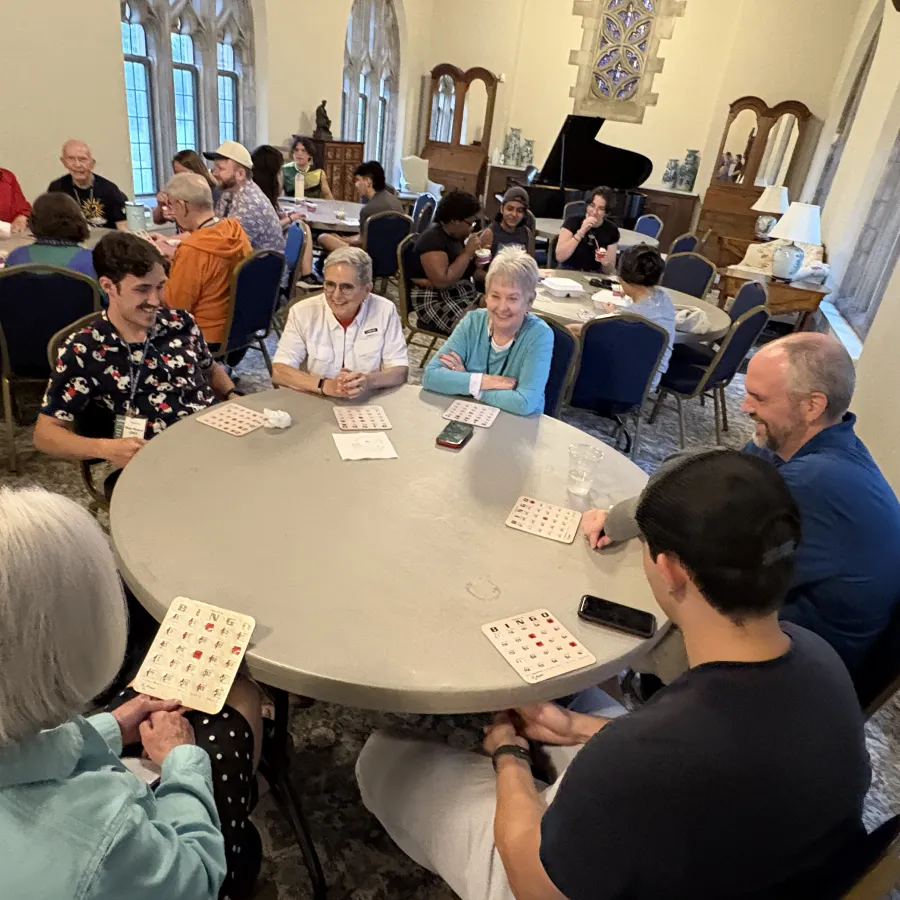 People of diverse ages play bingo around tables in a bright room with large windows and stone walls.