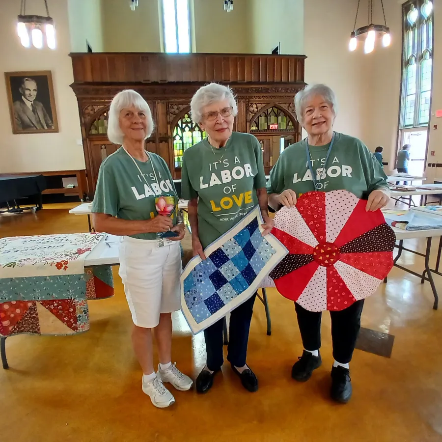 Three elderly women wearing matching labor of love shirts display handmade quilts inside a bright community hall.
