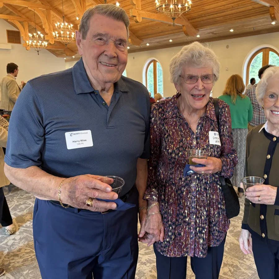 Three elderly people smiling and holding drinks at an indoor social event with wooden beams and arched windows.