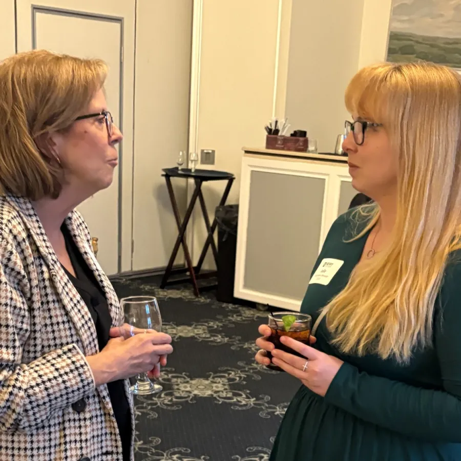 Two women conversing indoors at a formal event, one in green dress and the other in checkered blazer holding drinks.
