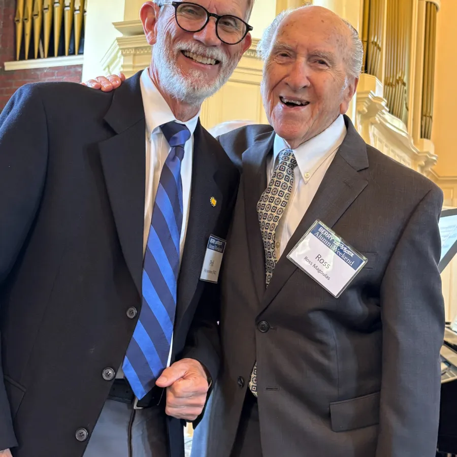 Two elderly men smiling and posing together at a formal event in suits with name badges indoors.