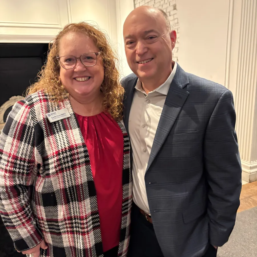 Smiling man in blue suit and woman in plaid jacket posing indoors in a formal setting with white walls.