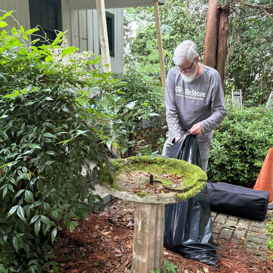Person wearing a gray Restore shirt gathers yard debris near a moss-covered birdbath in a leafy garden area.