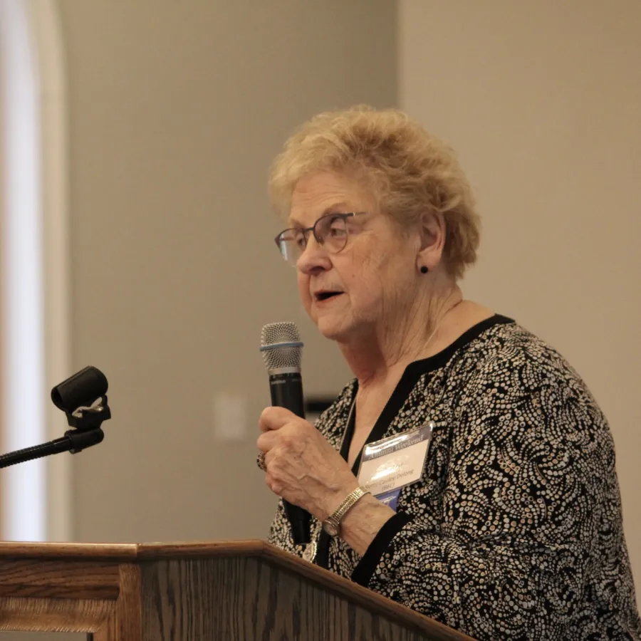Older woman in glasses speaking into microphone at wooden podium during indoor event